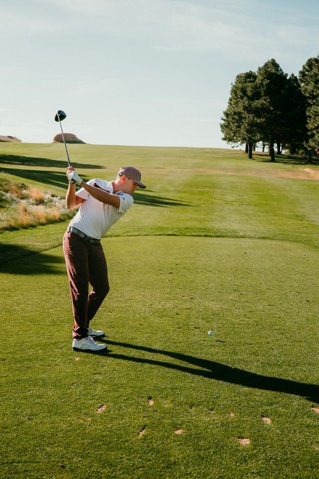 Golfer in white shirt and brown pants swinging a club on a green golf course under a blue sky.