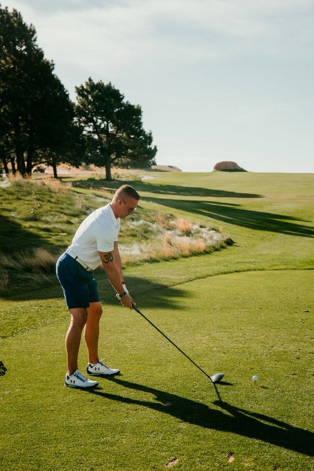 Man teeing off on a golf course. He wears a white shirt, blue shorts, and golf shoes on a sunny day.