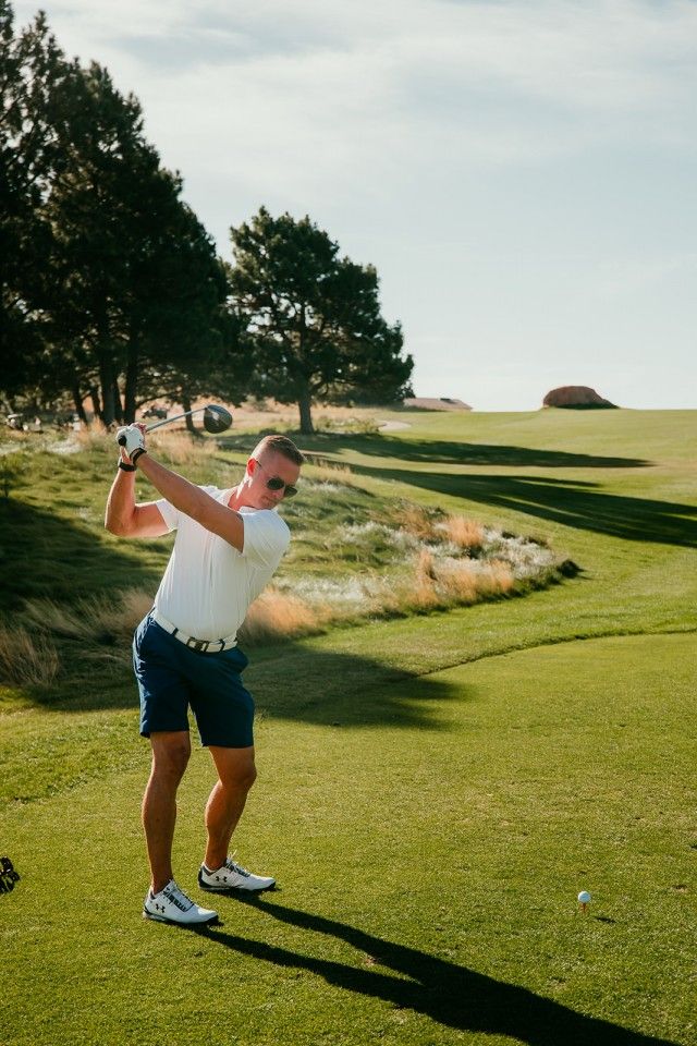 Golfer in white shirt and blue shorts swings club on a sunny golf course.