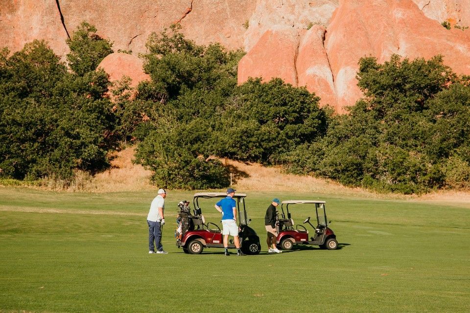 Three golfers near golf carts on a green course, reddish rock formations and trees in the background.