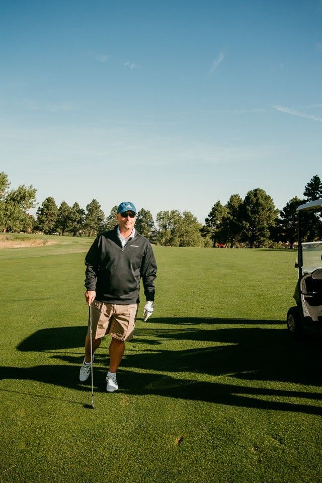 Man on golf course holding club, wearing a jacket, hat, shorts. Sunny day, golf cart visible.