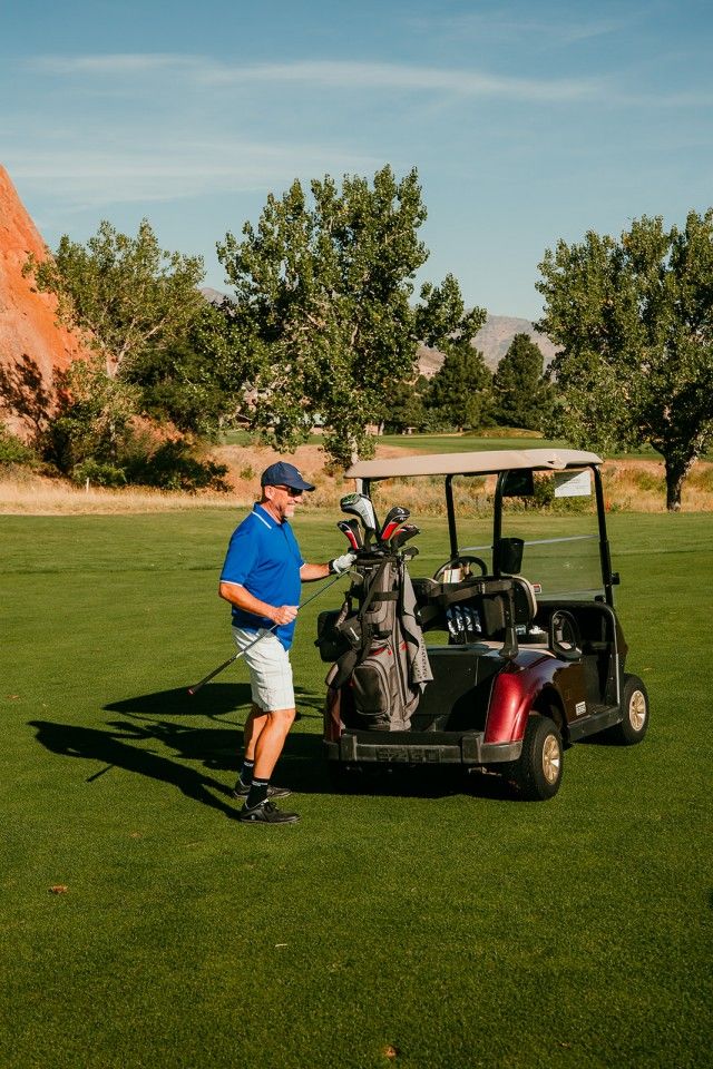 Man in blue shirt and shorts standing by a golf cart on a green golf course under a blue sky.