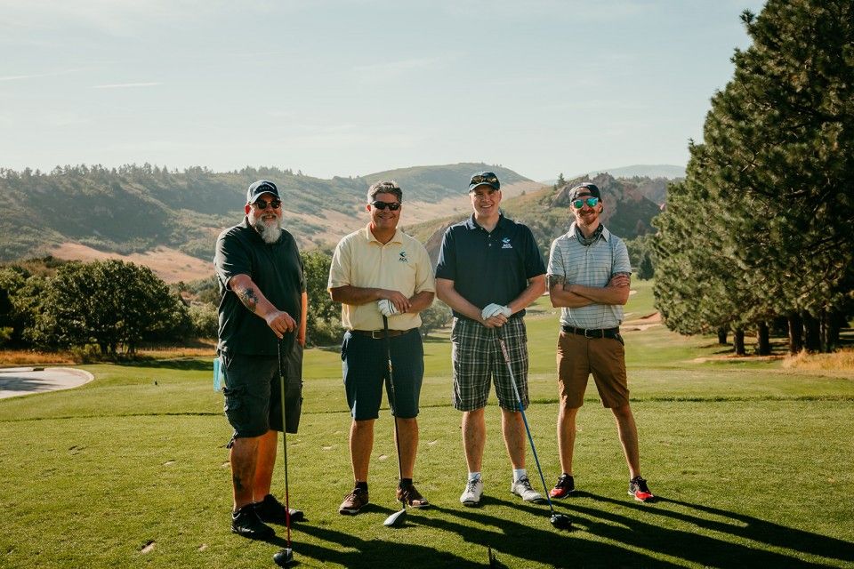 Four golfers standing on a green, holding clubs. Mountainous golf course backdrop, sunny day.