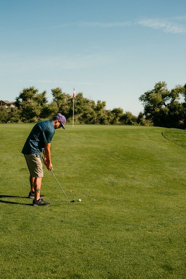 Man in blue shirt and purple cap playing golf on a green course under a blue sky.