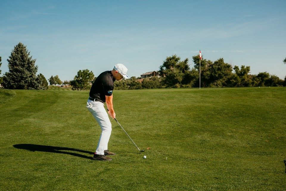 Golfer in white pants and black shirt swings golf club on green course with flag in background.