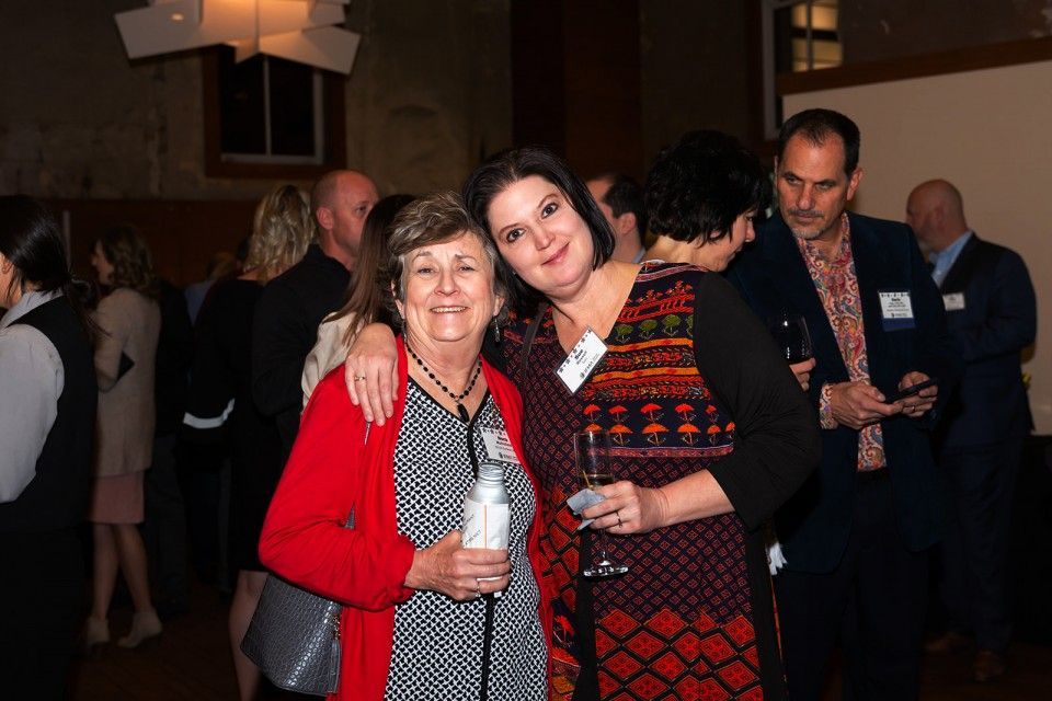 Two women smiling and posing at an event. One in a red jacket, the other with an arm around her, holding a drink.