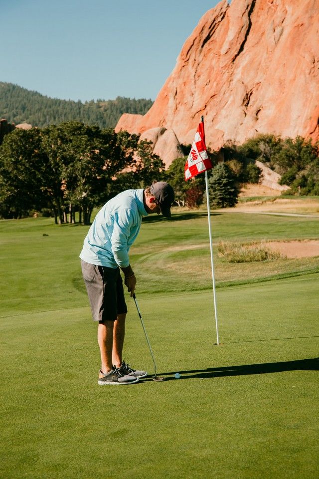 Golfer putting on green, with red rock formations and flag in the background.
