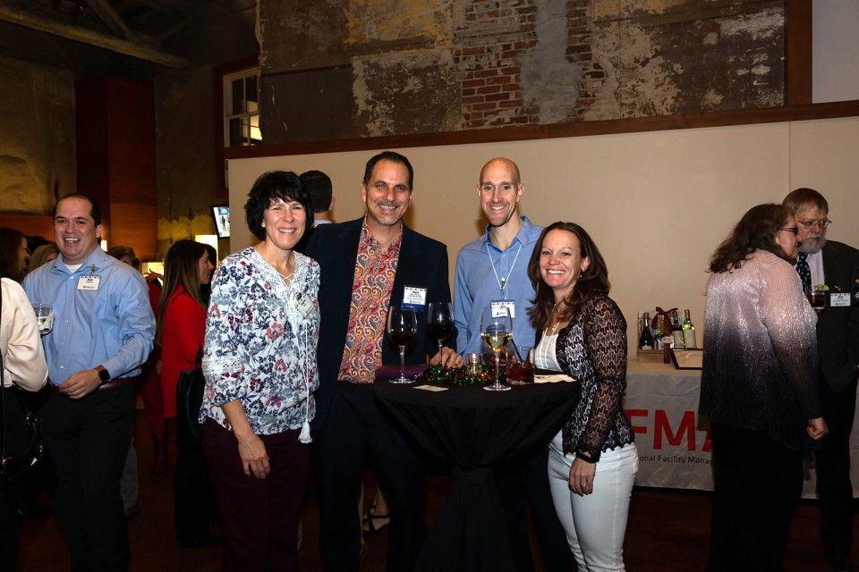 Four people at an event stand near a table with drinks; others are in the background.