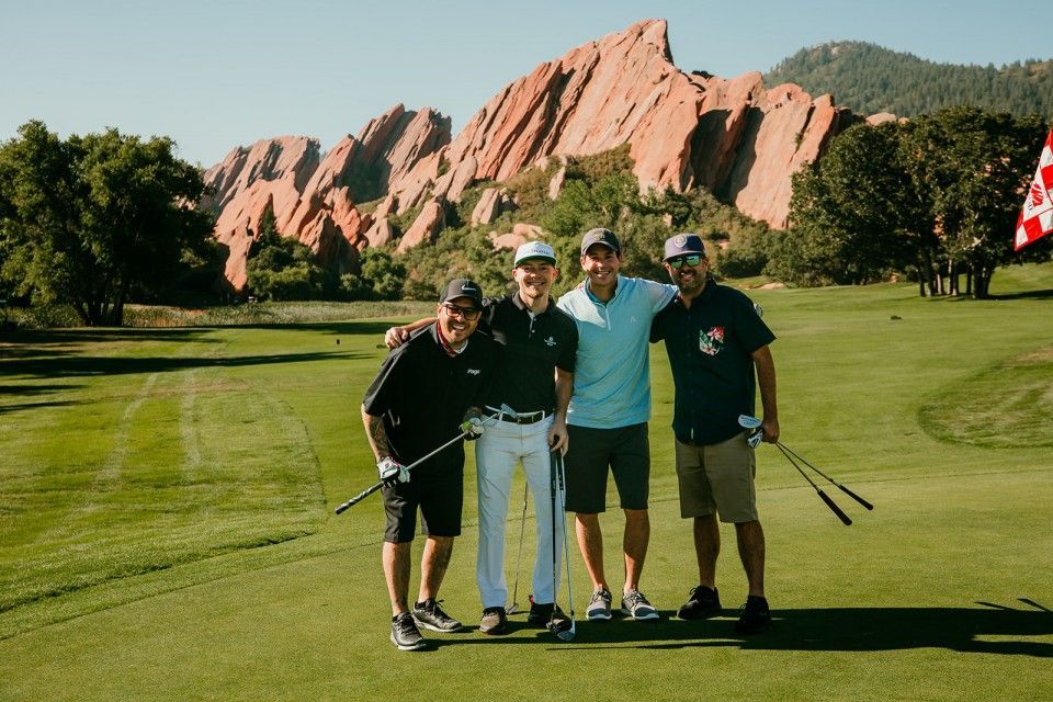 Four men pose on a golf course with red rock formations in the background. They are holding golf clubs and smiling.