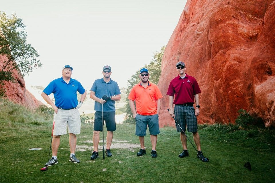 Four men in golf attire stand on a green, red rock backdrop.