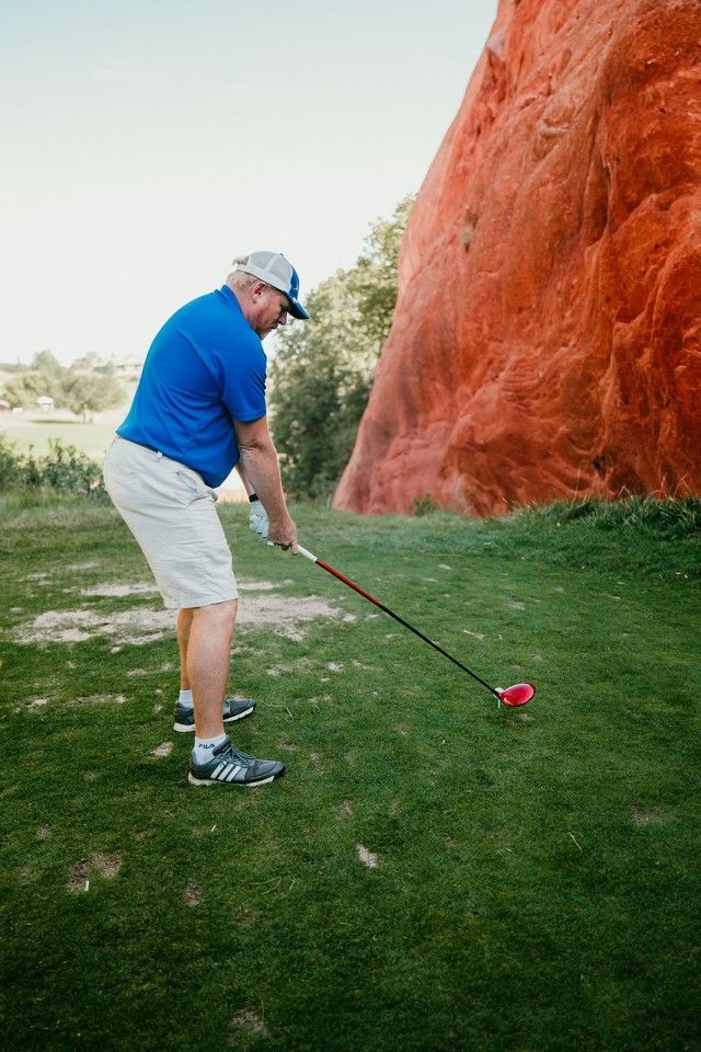 Man in blue shirt and white shorts golfing, holding a club on a green course, with a red rock formation in the background.