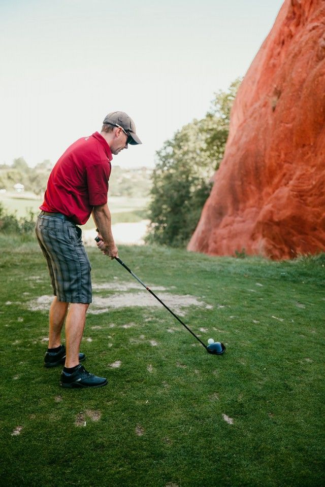 Man in red shirt and shorts teeing off on a golf course, with a large red rock formation in the background.