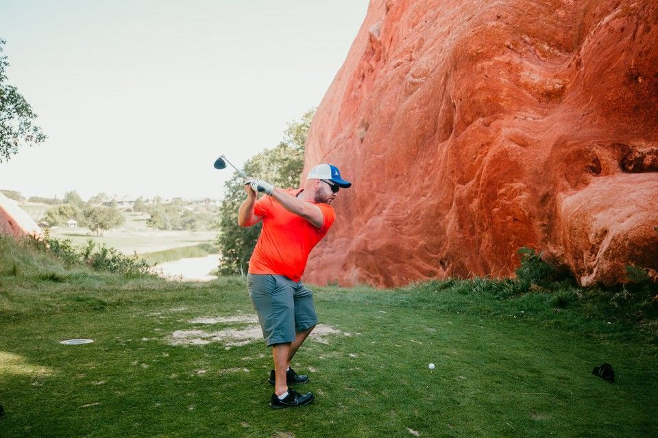 Man in orange shirt swings golf club on green, red rock formation in background.