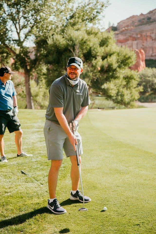 Man in gray shirt putting on a golf course; another man watches. Green grass and trees in background.