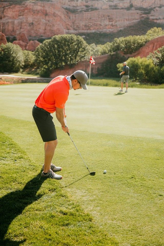Man putting a golf ball on a green, red flag in the background, another golfer further back.