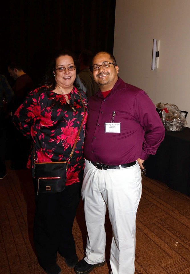 Two people smiling at the camera, indoors. Woman in black pants and floral shirt; man in white pants and purple shirt.