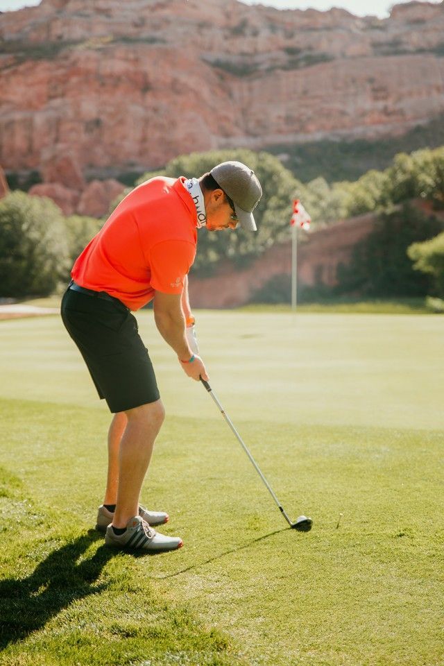 Golfer taking a shot on a green course with red rock formations in the background.