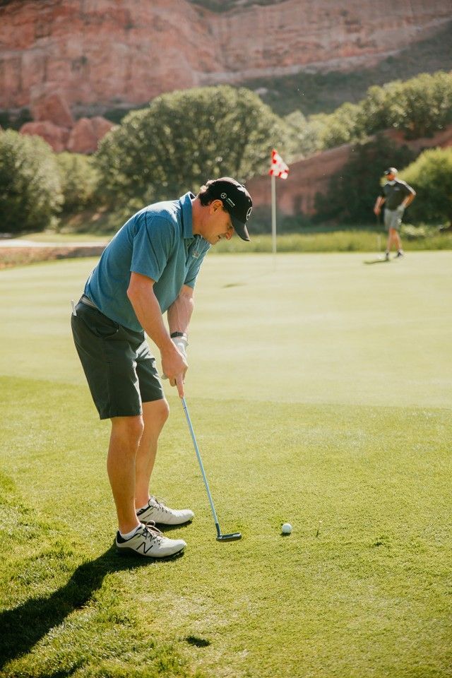 Man putting golf ball on a green, another golfer in the background, red rock cliffs.
