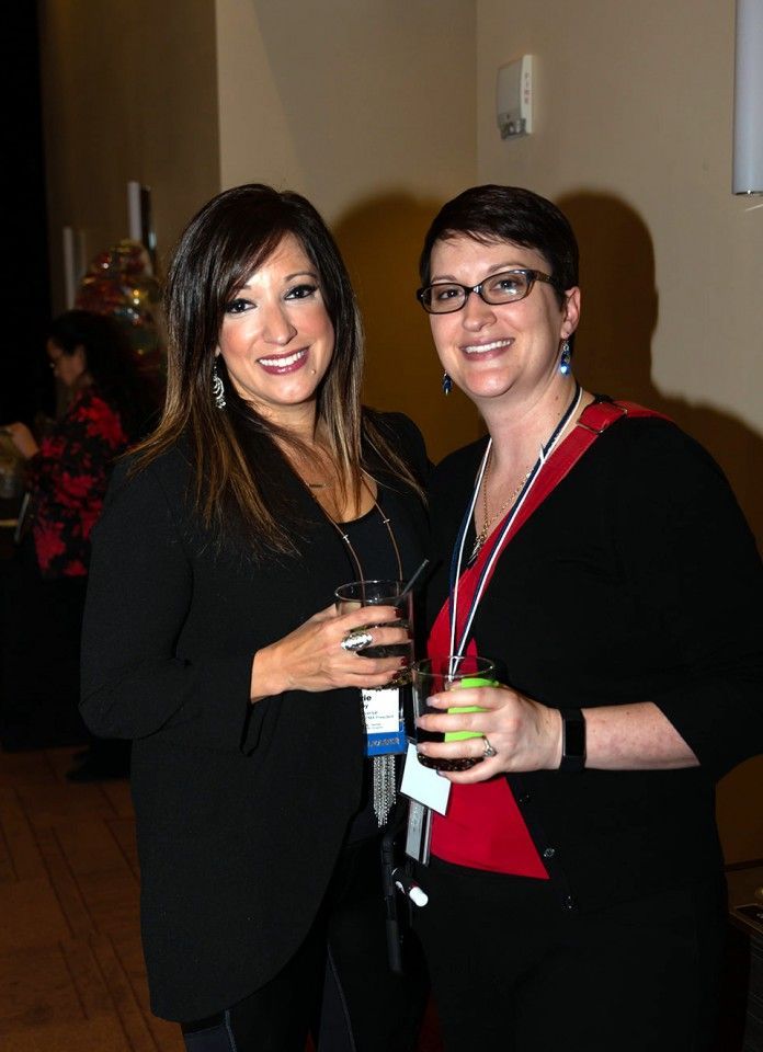 Two women smiling, holding drinks, indoors. One in a black blazer, the other with glasses and a red shirt.