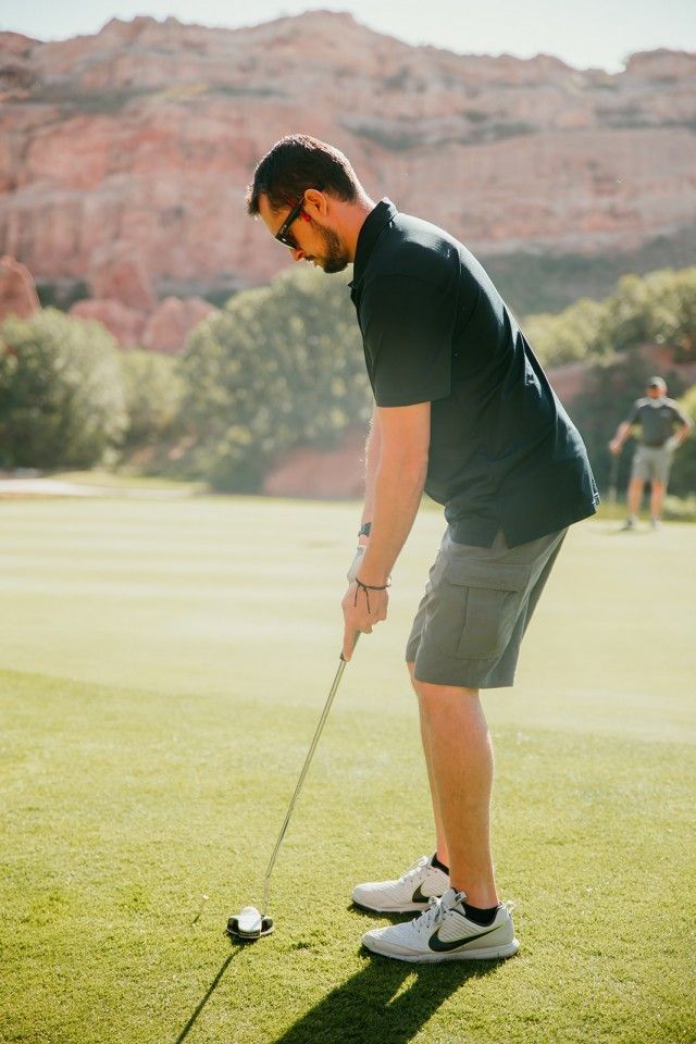 Man putting a golf ball on a green, wearing a navy polo and gray shorts; red rock backdrop.