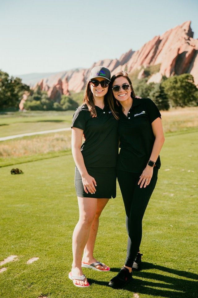 Two women in black polo shirts and sunglasses pose on a golf course with red rock formations in the background.