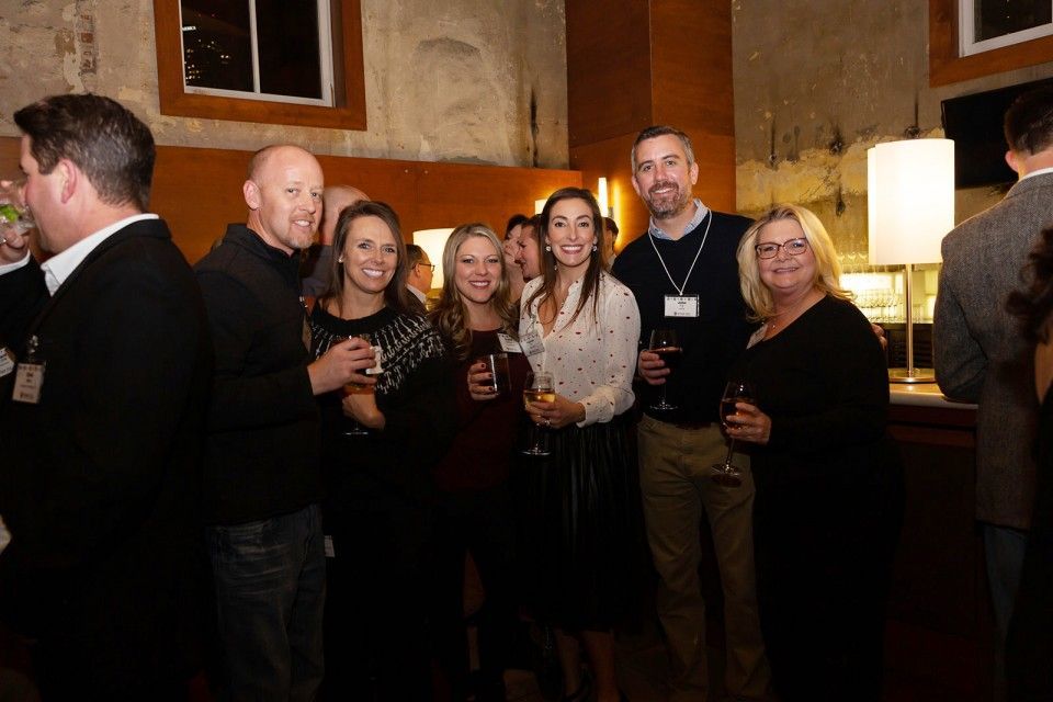 Group of people at an event, some holding drinks, smiling. Dark setting, neutral tones.