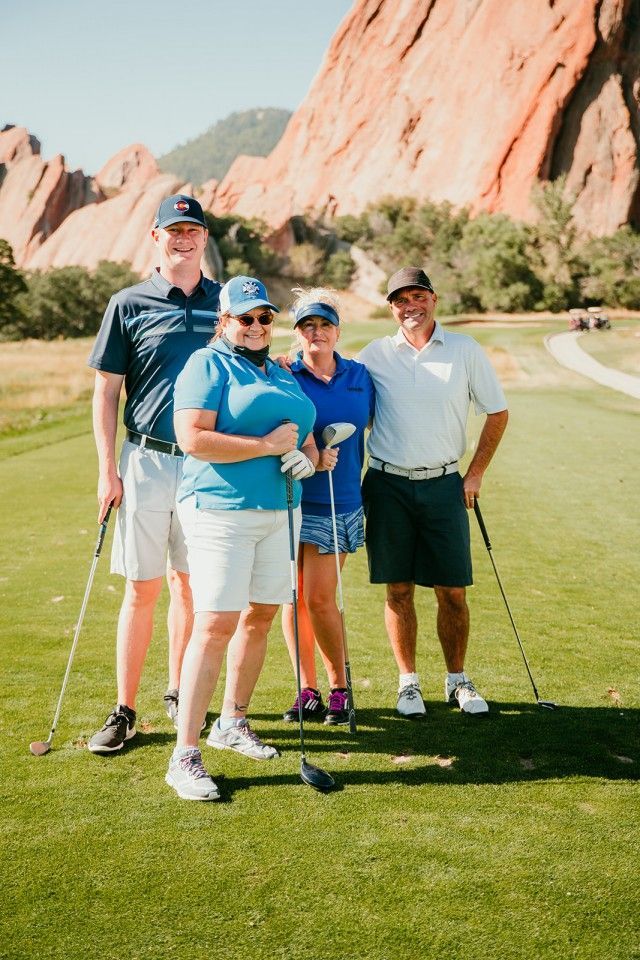 Four people on a golf course pose, wearing golf attire. Red rock formations are in the background.