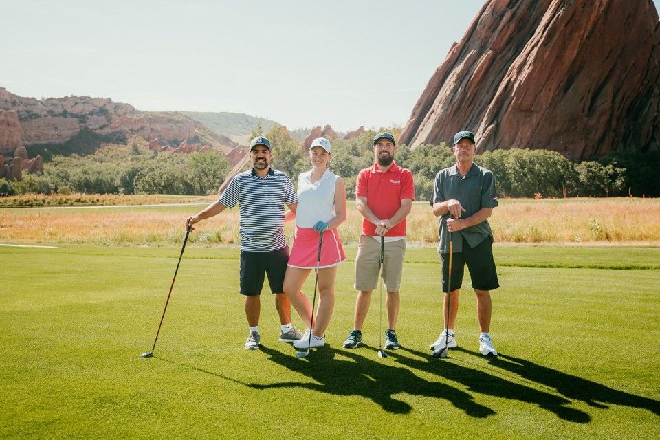 Four people on a golf course posing for a photo. Red rock formation in the background. Sunny, green grass.