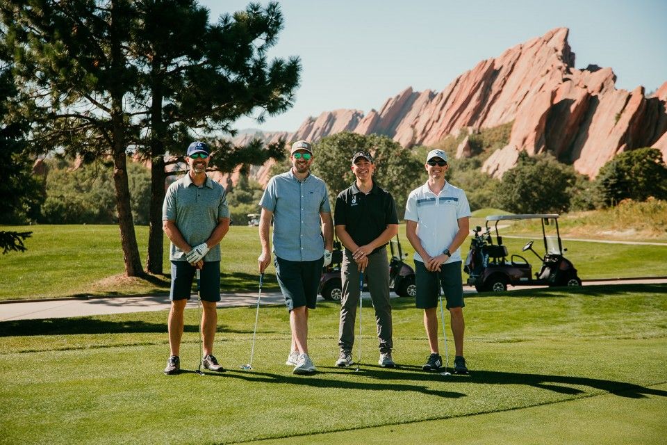 Four men stand on a golf course with golf clubs, red rock mountains in the background, blue sky.