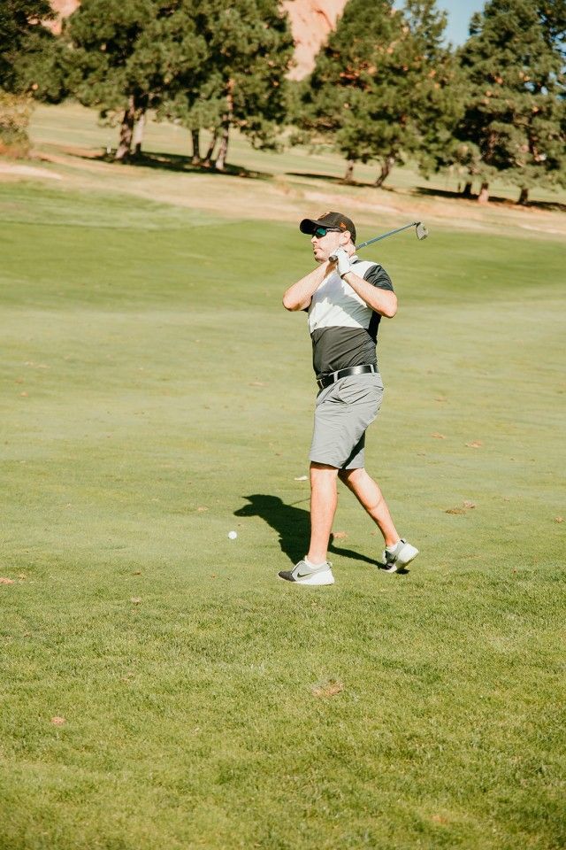 Man swinging golf club on green grass. Ball on the ground, trees in the background.