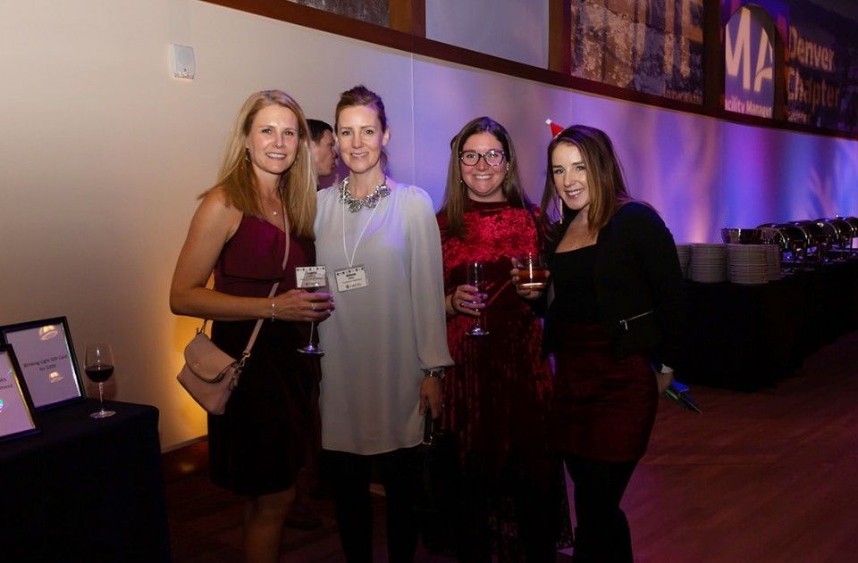 Four people at an event, smiling and holding drinks. Dark outfits, interior setting with food and WA sign.