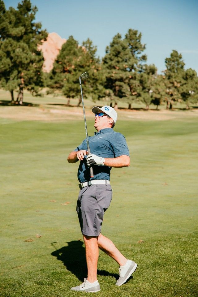 Golfer on a green golf course, raising club after swing, wearing shorts, hat, and shirt.