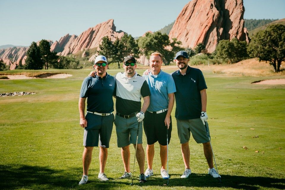 Four men pose on a golf course with red rock formations in the background. They wear golf attire.