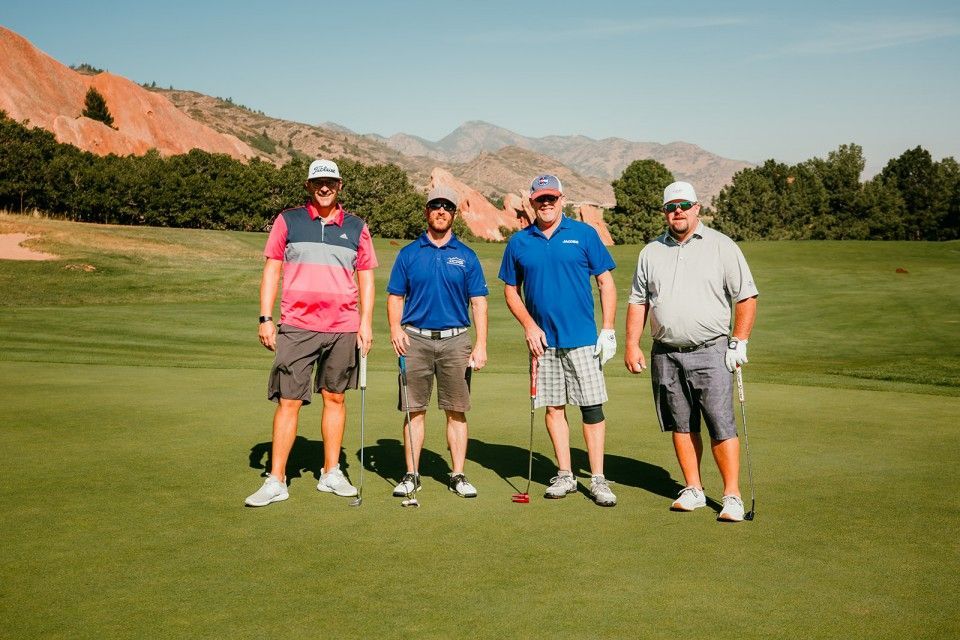 Four men on a green golf course, posing for photo. Mountains and red rock formations in background.