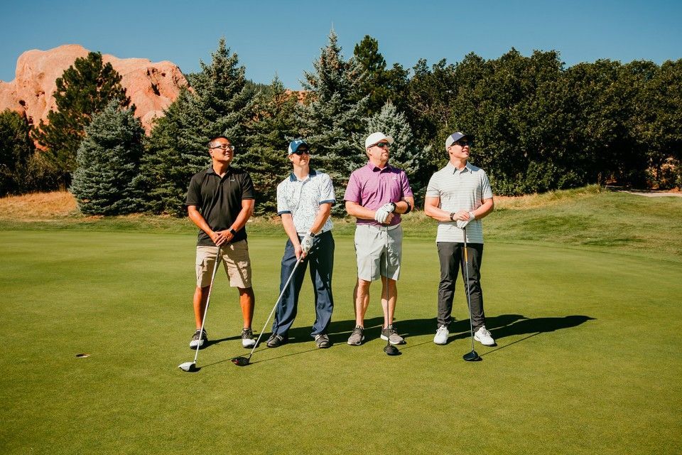 Four golfers on a green field with golf clubs. Trees and red rock formations in the background. Sunny day.