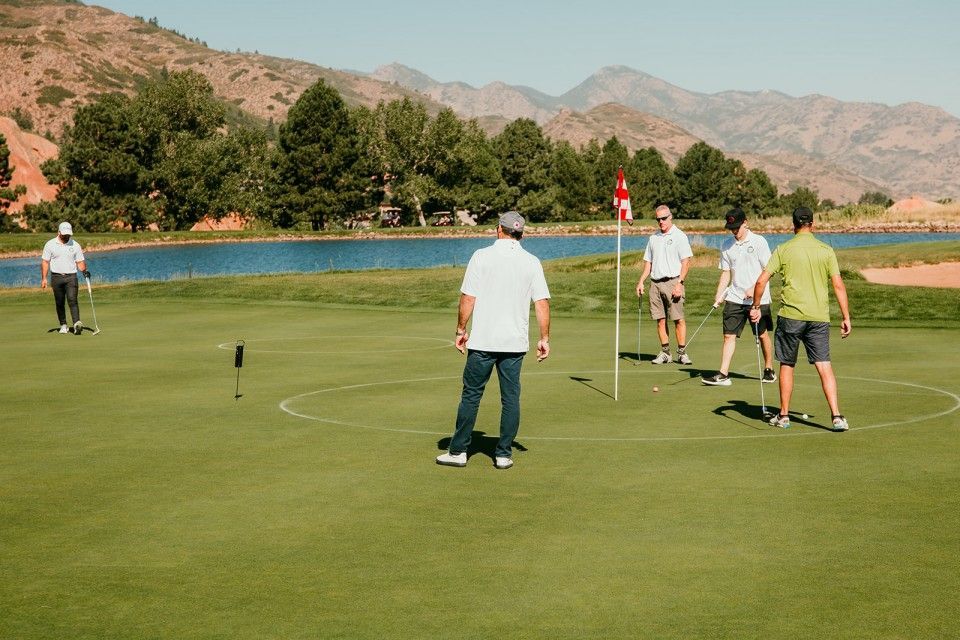 Men playing golf on a green, with a lake and mountains in the background.