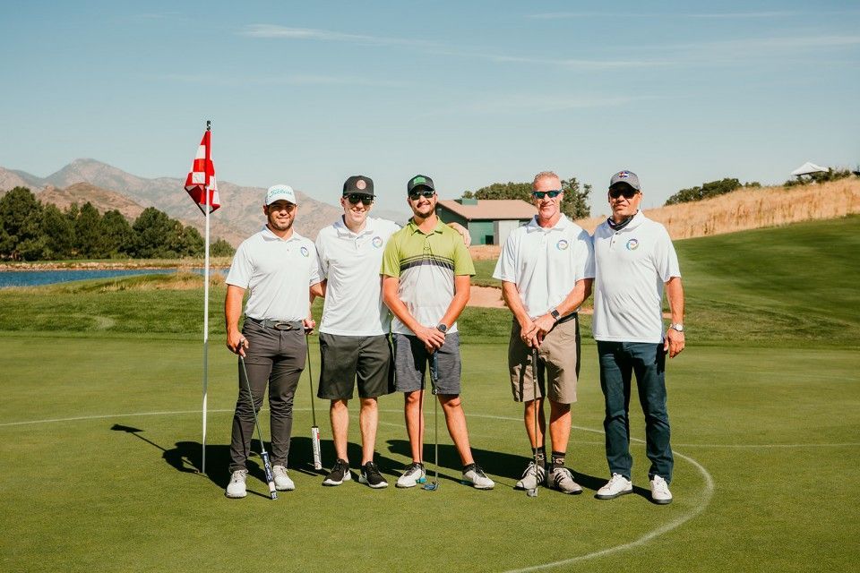 Five men pose on a golf course green; mountains in the background.