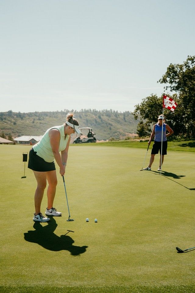Woman putts golf ball on green, another person stands by flag. Sunny day.