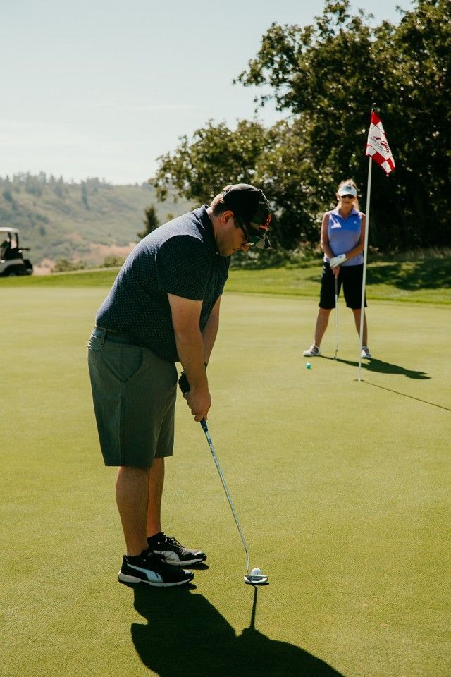 Man putting a golf ball on a green, woman holding the flag in background. Sunny day on a golf course.