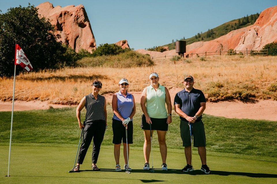 Four people on a golf course posing near the green with a flag in the background, red rock landscape.