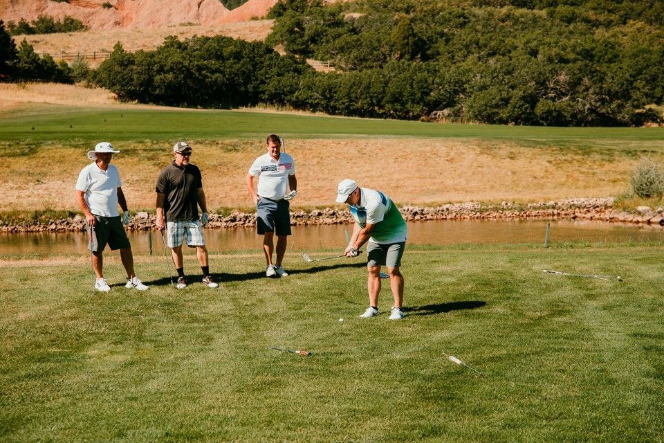 Four men on a golf course: one about to swing, others watching. Green grass, blue shorts, trees in background.