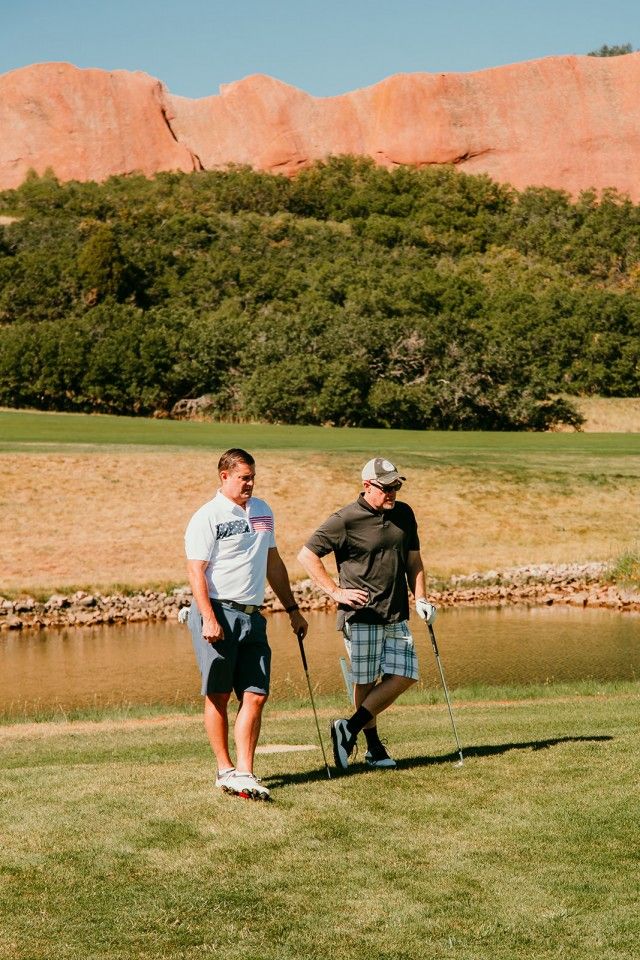 Two men on a golf course with red rock in the background, holding golf clubs. Sunny day.