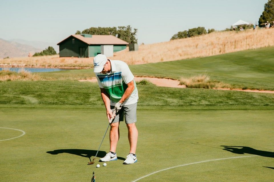 Man putting golf ball on a green, sunny day. He wears a white hat and shirt, gray shorts, and white sneakers.