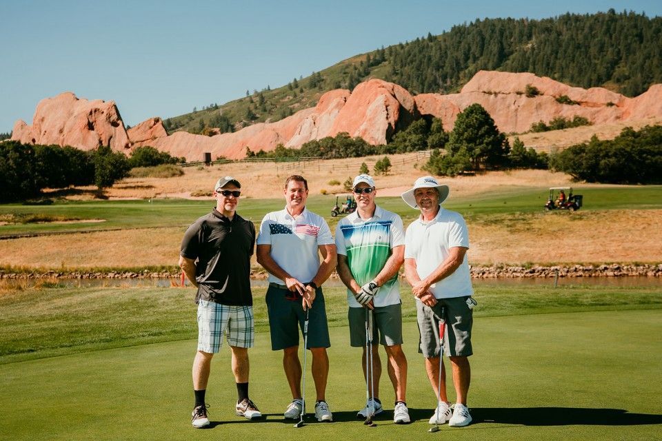 Four men with golf clubs on a green course, red rock mountains in the background, sunny day.