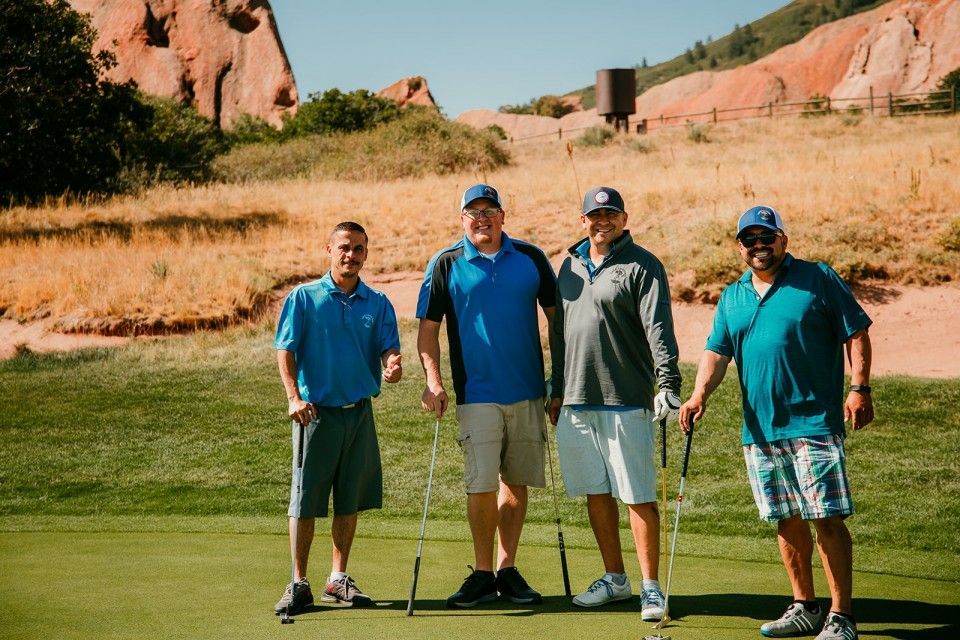 Four men stand on a golf course, holding putters. Red rock mountains in background.