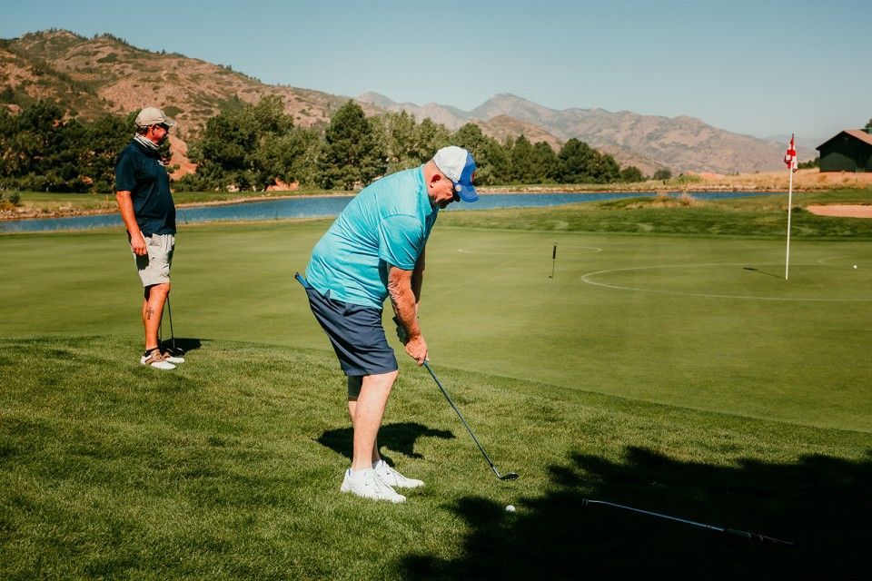 Two men playing golf on a green course; one tees off, the other watches near a water feature and mountains.