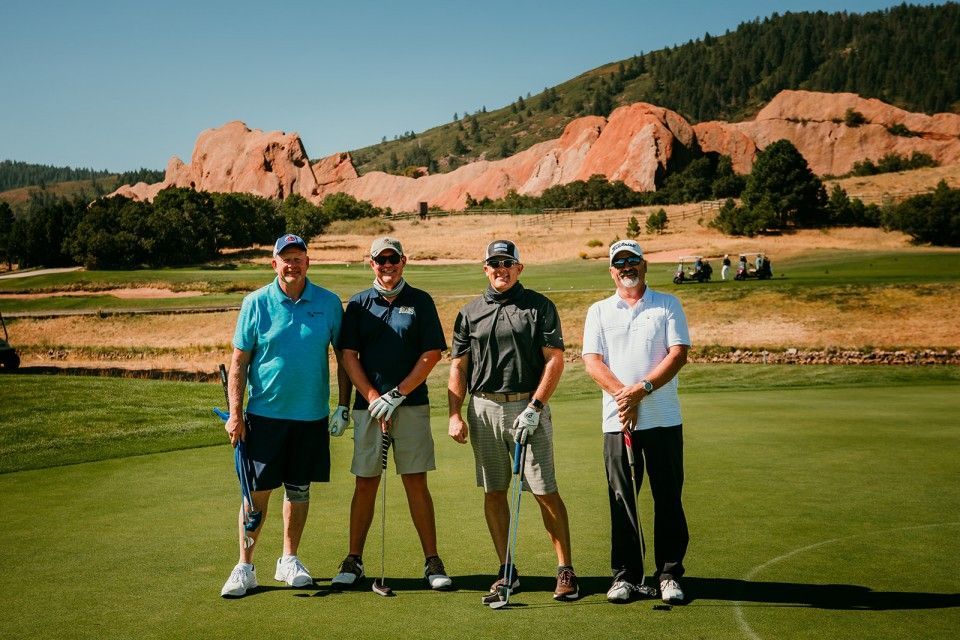 Four men pose with golf clubs on a green, red rock formation in background. Sunny day.