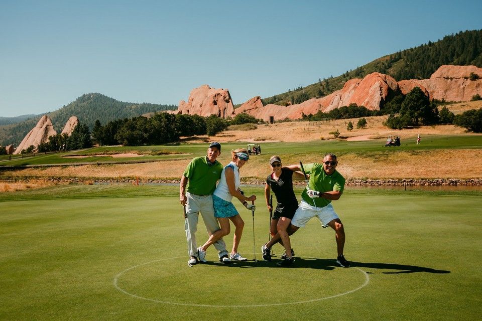 Four golfers pose on green with golf clubs, red rock mountains in background.