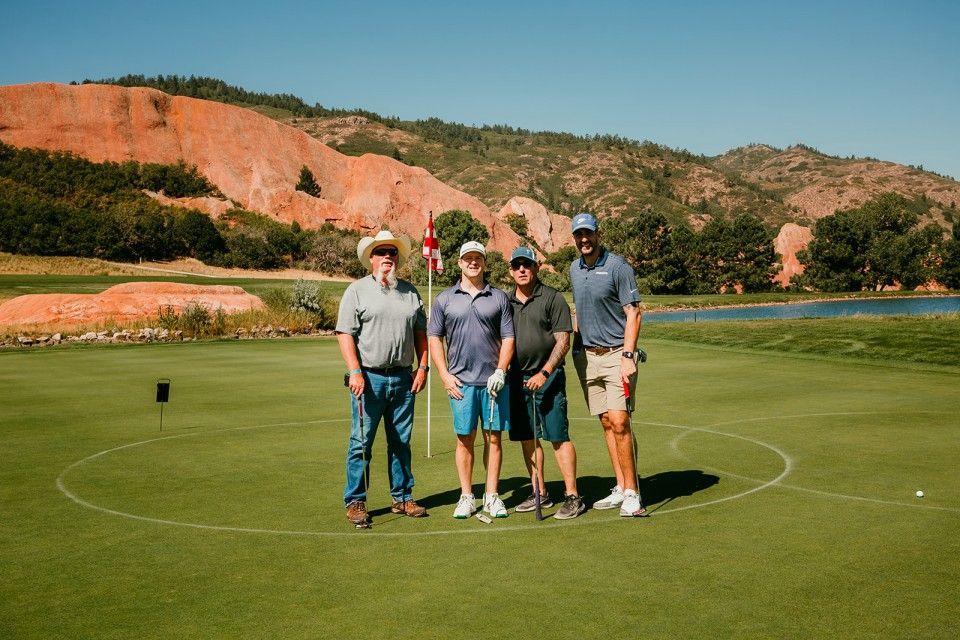 Four golfers standing on a green, red rock mountains in the background, sunny day.