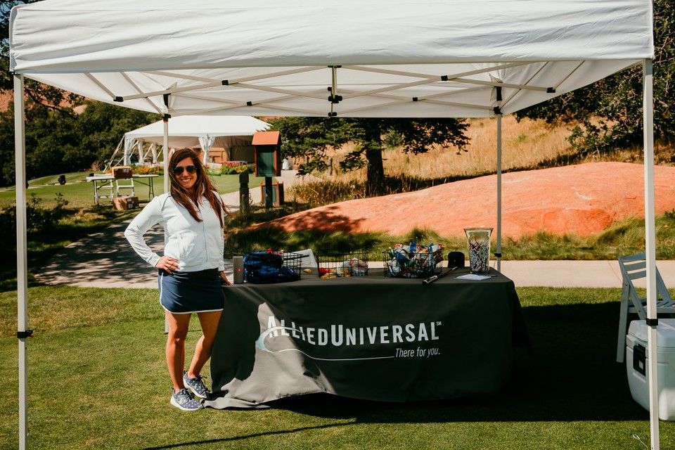 Woman stands by a branded tent at a golf course. She wears athletic attire and smiles. The tent displays items.